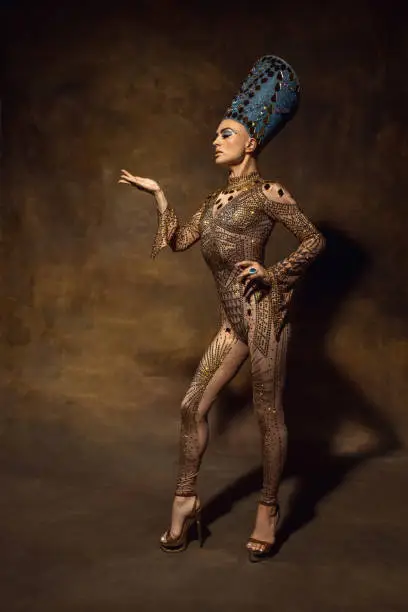 Young woman in image of Nefertiti in art performance isolated on brown vintage background. Beautiful female model like famous queen of Egypt. Retro style, comparison of eras concept.