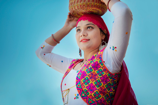 Low angle image of beautiful traditional young woman carrying wooden basket on her head and standing against blue sky and looking away with smile.