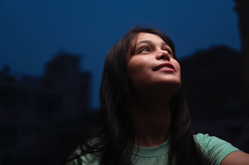Low angle portrait of Asian/Indian serene young woman looking up with smile and contemplating at night.