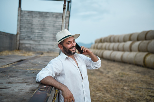Smiling Bearded Modern Farmer Calling Workers To Help Load Hay Bales On Caravan