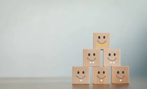 Photo of A wooden blog on customer service assessment concepts and satisfaction surveys. The customer selects a smiley face icon and a star symbol lined up on the upper wooden block of the wooden cube on the table.