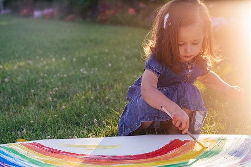 Little girl 2-4 years old paints rainbow and sun on large sheet of paper sitting on green lawn in sunlight