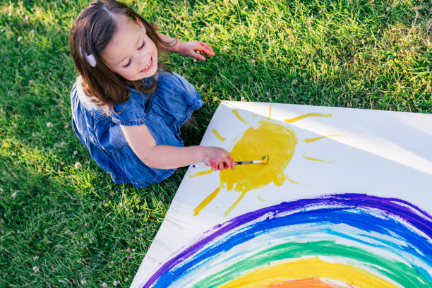 petite fille de 2-4 ans peint arc-en-ciel et soleil sur une grande feuille de papier, assis sur la pelouse verte - child caucasian little girls 3 4 years photos et images de collection