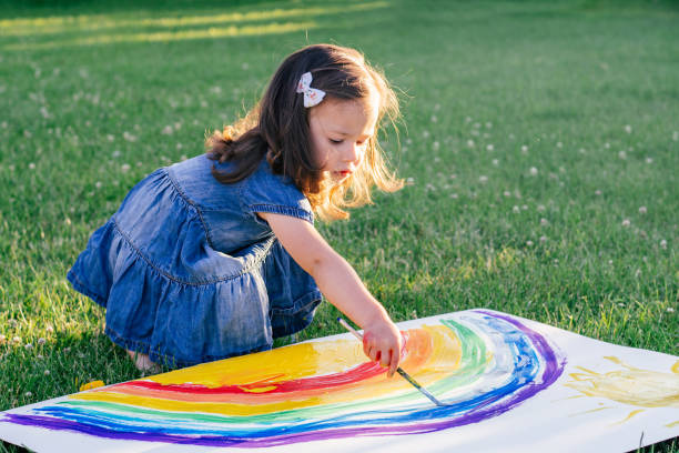 dziewczynka w wieku 2-4 lat maluje tęczę i słońce na dużej kartce papieru, siedząc na zielonym trawniku - child caucasian little girls 3 4 years zdjęcia i obrazy z banku zdjęć