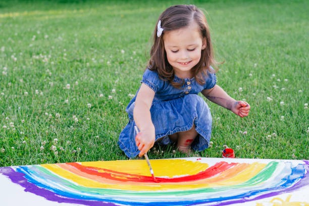 petite fille de 2-4 ans peint arc-en-ciel et soleil sur une grande feuille de papier, assis sur la pelouse verte - child caucasian little girls 3 4 years photos et images de collection