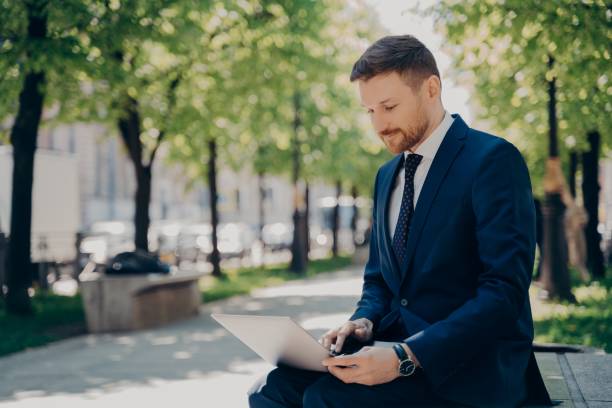 istock Young businessman working on laptop while sitting on bench in city park 1328705775