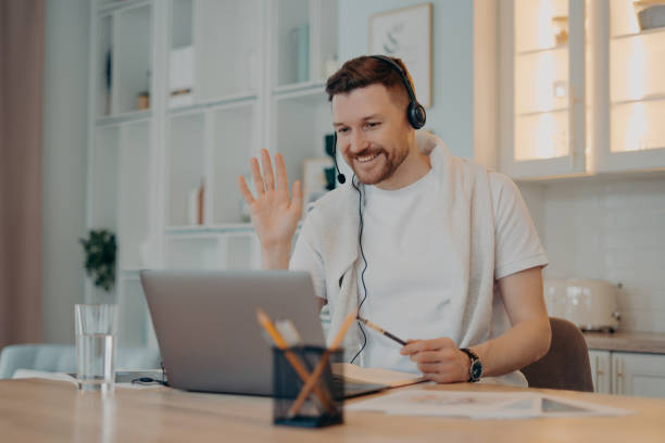 istock E learning concept. Happy bearded man student has video conference studies online waves palm in laptop display greets teacher works distantly uses headset makes notes in diary poses over home interior 1328708203