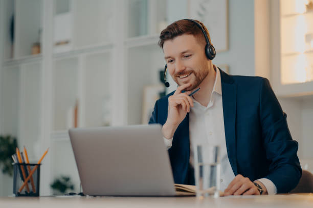 istock Professional male operator works in callcenter uses headset and laptop computer talks on video call or virtual webcam event wears formal clothes poses against cozy interior has online meeting 1328708382