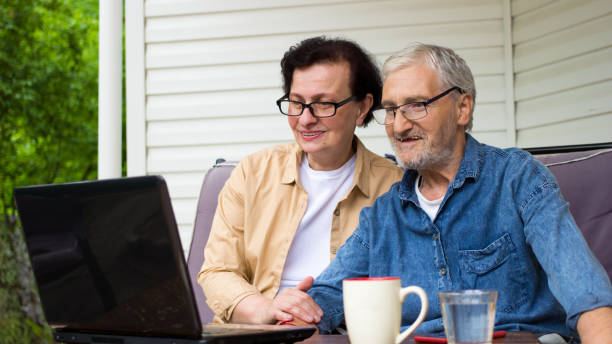 ノートパソコンの画面を見て微笑むシニアカップルは、屋外のホームテラスのソファに座っています。技術を使用して成熟し、引退した男女。高齢者家族読書ニュース、オンラインショッピ� - grandparent senior adult child reading ストックフォトと画像