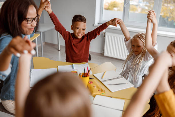 istock Smiling kids sitting at the table and raising their hands up 1328727395