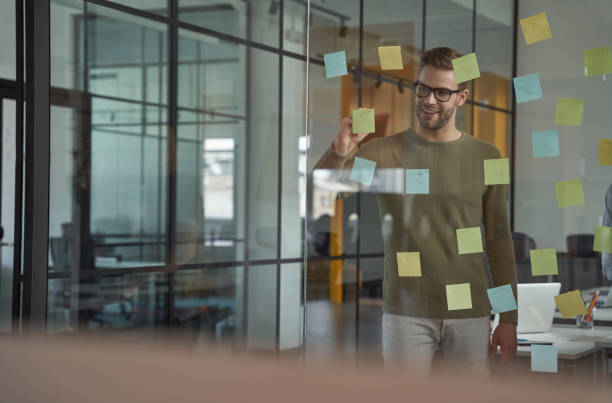 istock Planning new project. Young caucasian man, office worker wearing glasses writing something on sticky notes on a glass board and smiling while working in the modern office 1328733812