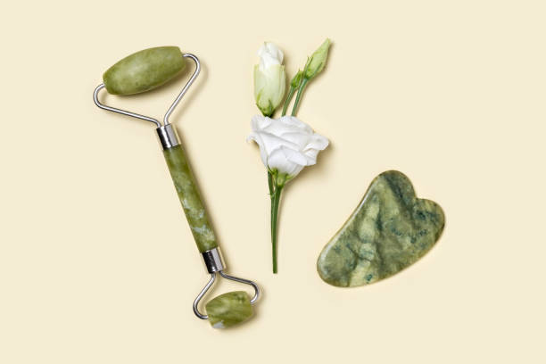 rullo di giada verde e pietra gua sha per massaggio facciale e tenero fiore bianco eustoma su sfondo giallo pastello. dispositivo a rulli per viso di bellezza domestico per il trattamento delle rughe anti età, il sollevamento o la tonizzazione - shiatsu beauty relaxation care foto e immagini stock