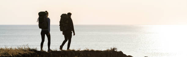 海の近くの山頂を歩くバックパックを持つ2人の旅行者 - climbing men sea cliff ストックフォトと画像