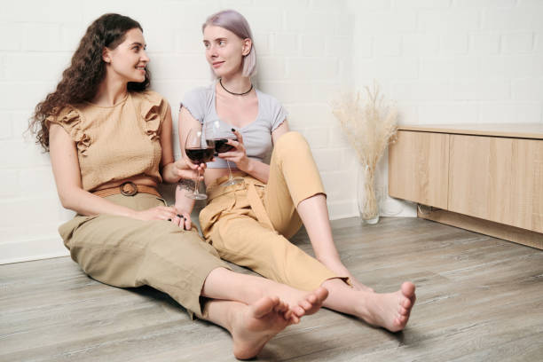 istock Happy young Caucasian lesbians sitting on floor and clinking wineglasses while drinking wine at home 1328757053