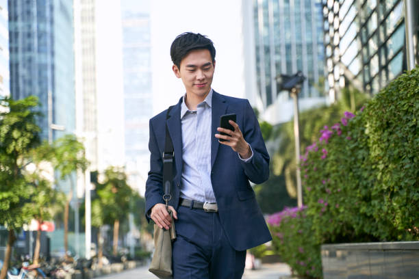 young asian business man office worker looking at mobile phone while walking on street young asian businessman looking at messages on cellphone while walking in the street in downtown of modern city shenzhen technology stock pictures, royalty-free photos & images
