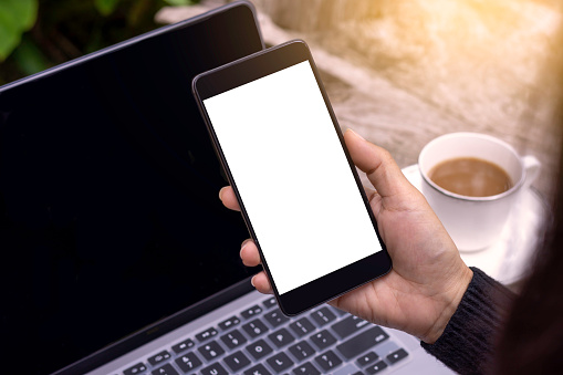 Mockup business person hand holding and showing mobile smart phone with laptop computer on the wooden table at outdoors cafe or coffee shop.