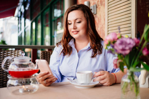 une femme séduisante en robe bleue boit du thé café et des sms dans un smartphone - restaurant business person setting the table clothing photos et images de collection