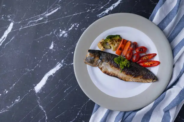 steak pacific mackerel grilled with sidedish broccoli, carrot, tomato, chilli and parsley on ceramic dish, marble table, dark black background, foodstyling concept, home cooking