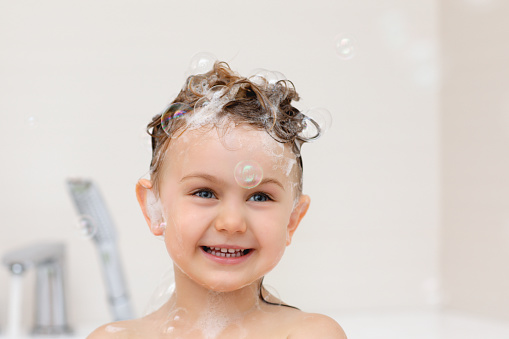 Cute happy smiling kid with white shampoo foam and bubbles on head taking bath. Child's health care and hygiene concept. Copy space