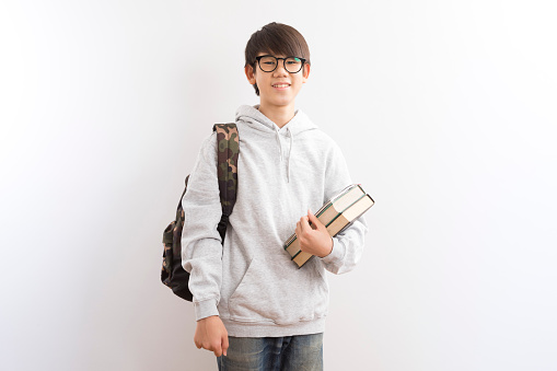 Asian teen boy students wearing hoodie and glasses carrying a backpack and books standing on white background.