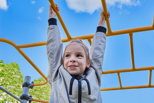 Happy cheerful and smiling Little girl 7-9 years playing on a playground, hanging walk along the monkey bars. Against the background of a blue sky with clouds on a sunny day in spring, summer. High quality photo