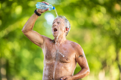 Old man with no shirt pours water from a bottle over his head and face outdoors on a hot summer day.