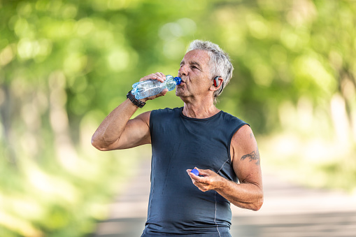 Handsome elderly man rehydrates after a run outdoors in the nature by drinking water from a bottle.