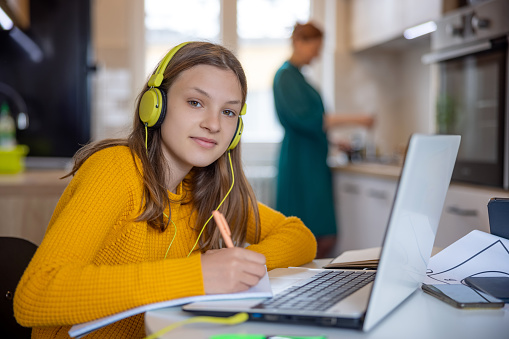 Teenage girl with headphones sitting at the table at home and writing in her notebook, looking at camera, her mother in the kitchen in blurred background