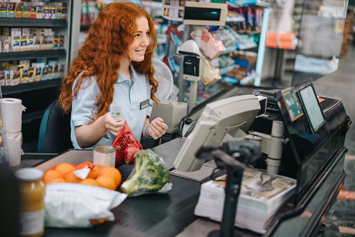 Young sales clerk sitting by cash register in supermarket and serving shoppers. Woman cashier scanning grocery products at checkout.