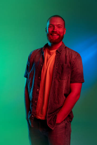 porträt eines jungen gutaussehenden lächelnden kaukasischen mannes mit händen in taschen, isoliert auf grünem hintergrund in blauem neonlicht. - neon fotos stock-fotos und bilder