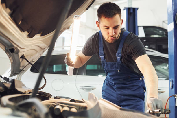 istock Uses lighting equipment. Man in work uniform repairs white automobile indoors. Conception of automobile service 1328820660