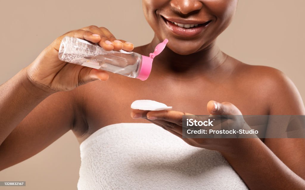 Beauty Routine. Closeup Of Black Female Using Micellar Water For Makeup Removing Beauty Routine. Closeup Of Black Smiling Female Using Micellar Water And Cotton Pad For Makeup Removing, African American Woman Cleansing Skin After Bath, Standing Over Beige Background, Cropped Removing Make-Up Stock Photo