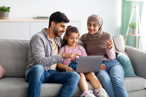 Modern muslim parents teaching their daughter using laptop computer, sitting together on sofa at home. Young arab man, child and woman in hijab staying home