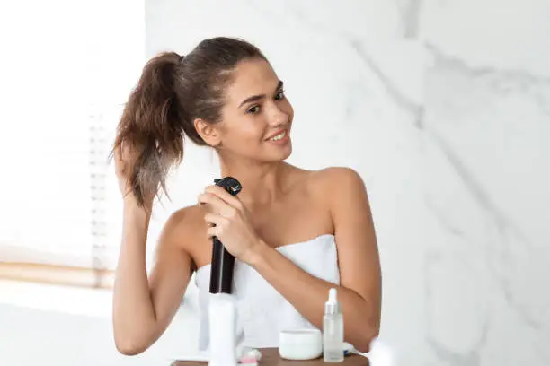 Haircare And Hairstyling. Cheerful Woman Spraying Hairspray On Hair For Repair, Making Ponytail Standing Near Mirror In Bathroom Indoor. Hair Care Cosmetics Advertisement Concept