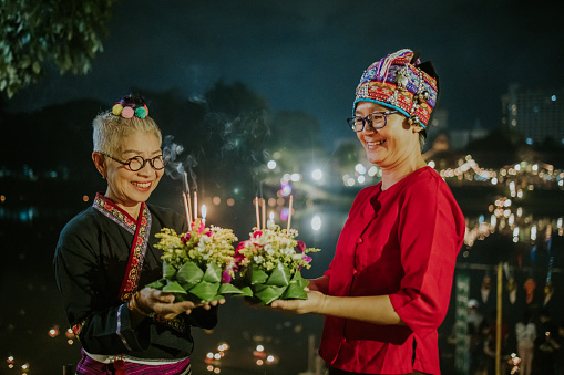 Two hipsters delightful Thai senior females in Thai local clothing look at Kratong with smiling and make a wish with  Thai local Kratong flower floating basket for a healthy and peaceful life together in Loy Kratong festival at night.
