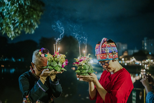 Loy Krathong festival in Chiangmai, Thailand, Two Thai senior women in Thai local clothing believe in the river spirit and make a wish with  Thai local Kratong flower floating basket for a healthy and peaceful life in Loy Kratong festival at night.