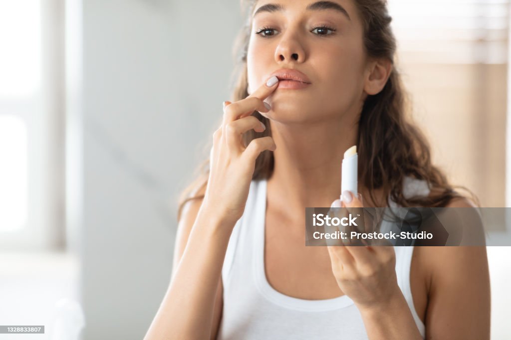Young Lady Applying Lip Balm Moisturizing Skin In Bathroom Indoors Young Lady Applying Lip Balm Moisturizing Skin Caring For Herself Standing In Modern Bathroom Indoors. Lips Skincare Concept, Beauty Routine. Cropped Shot, Selective Focus On Lipstick Lip Balm Stock Photo
