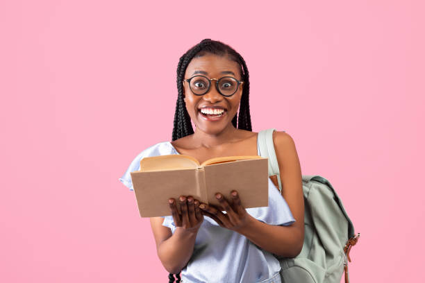 ピンクの背景にオープンブックを保持バックパックを持つ興奮した若いアフリカ系アメリカ人の女子学生 - scholarship holder ストックフォトと画像