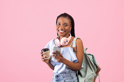 Online education. Young black woman with backpack, headphones, mobile device and takeaway coffee posing on pink studio background. Young female college student with afro bunches ready for school