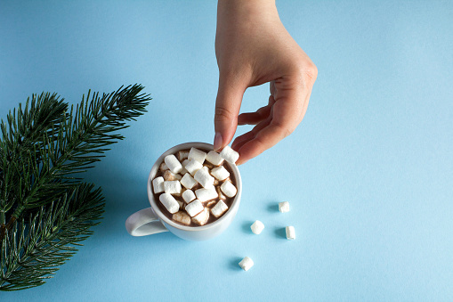 Hot chocolate with marshmallows in the white cup  and women hand  on the blue  background. Copy space. Close-up.