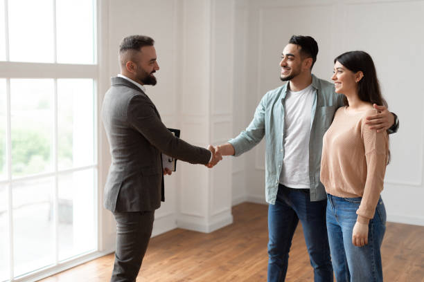 Millennial Couple Buying New Apartment, Shaking Hands With Real Estate Agent Successful Deal Concept. Portrait Of Young Family Buying New Apartment, Husband Embracing Wife Shaking Hands With Smiling Property Agent. Advisor In Suit Selling Flat To Happy Millennial Buyers estate agent stock pictures, royalty-free photos & images