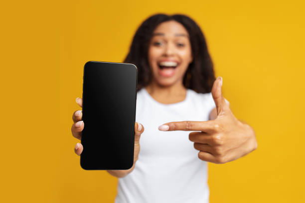 istock Cool mobile app. Overjoyed african american lady pointing finger at blank smartphone screen over yellow background 1328835580