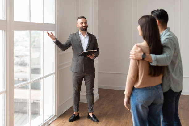 Estate Agent In Suit Showing Buyers New Empty Apartment Look Here. Professional House Agent Seller In Suit Showing New Apartment To Young Couple, Pointing At Beautiful View From Floor to Ceiling French Windows To Embracing Future Homeowners. Purchase exploration stock pictures, royalty-free photos & images