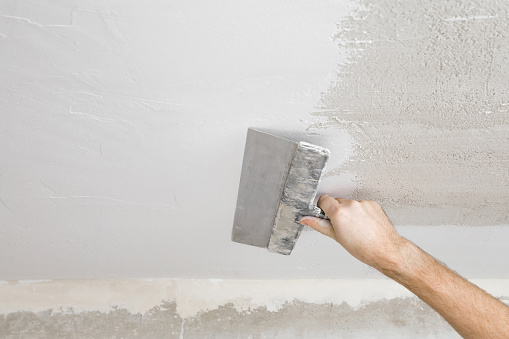 Young adult man hand using spatula and plastering ceiling with white fresh finishing putty. Closeup. Repair work of home. View from below.