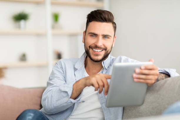 istock Handsome young man holding digital tablet and smiling at camera, sitting on couch at home, free space 1328839732
