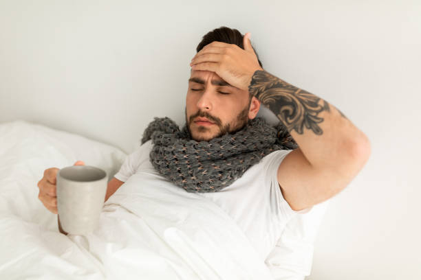 istock Feeling bad. Sick guy having fever, drinking hot healing tea and touching his forehead, lying in bed, empty space 1328840195