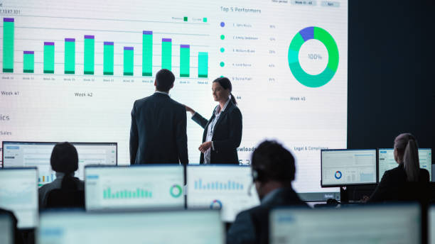 Two Traders Having a Meeting in a Modern Monitoring Office with Analytics Feed on a Big Digital Screen. Monitoring Room with Brokers and Finance Specialists Sit in Front of Computers. Colleagues Talk. dos comerciantes que tienen una reunión en una oficina de monitoreo moderna con analytics feed en una gran pantalla digital. sala de monitoreo con corredores y especialistas en finanzas se sientan frente a las computadoras. los colegas hablan.