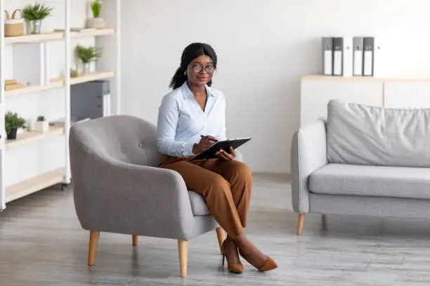 Photo of Full length portrait of friendly black female psychologist with clipboard sitting in armchair at office, copy space
