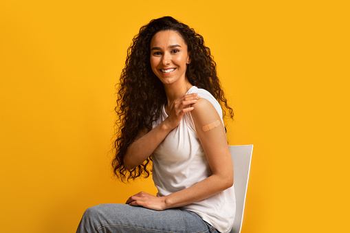 Portrait Of Smiling Young Woman Showing Arm After Coronavirus Vaccination, Happy Brunette Female Had Covid Vaccine Injection, Sitting On Chair With Rolled Up Sleeve Over Yellow Background, Copy Space