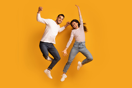 Cheerful Active Arab Couple Jumping In Air Over Yellow Background, Joyful Emotional Middle Eastern Man And Woman Having Fun Together, Raising Hands And Exclaiming With Excitement, Full Length